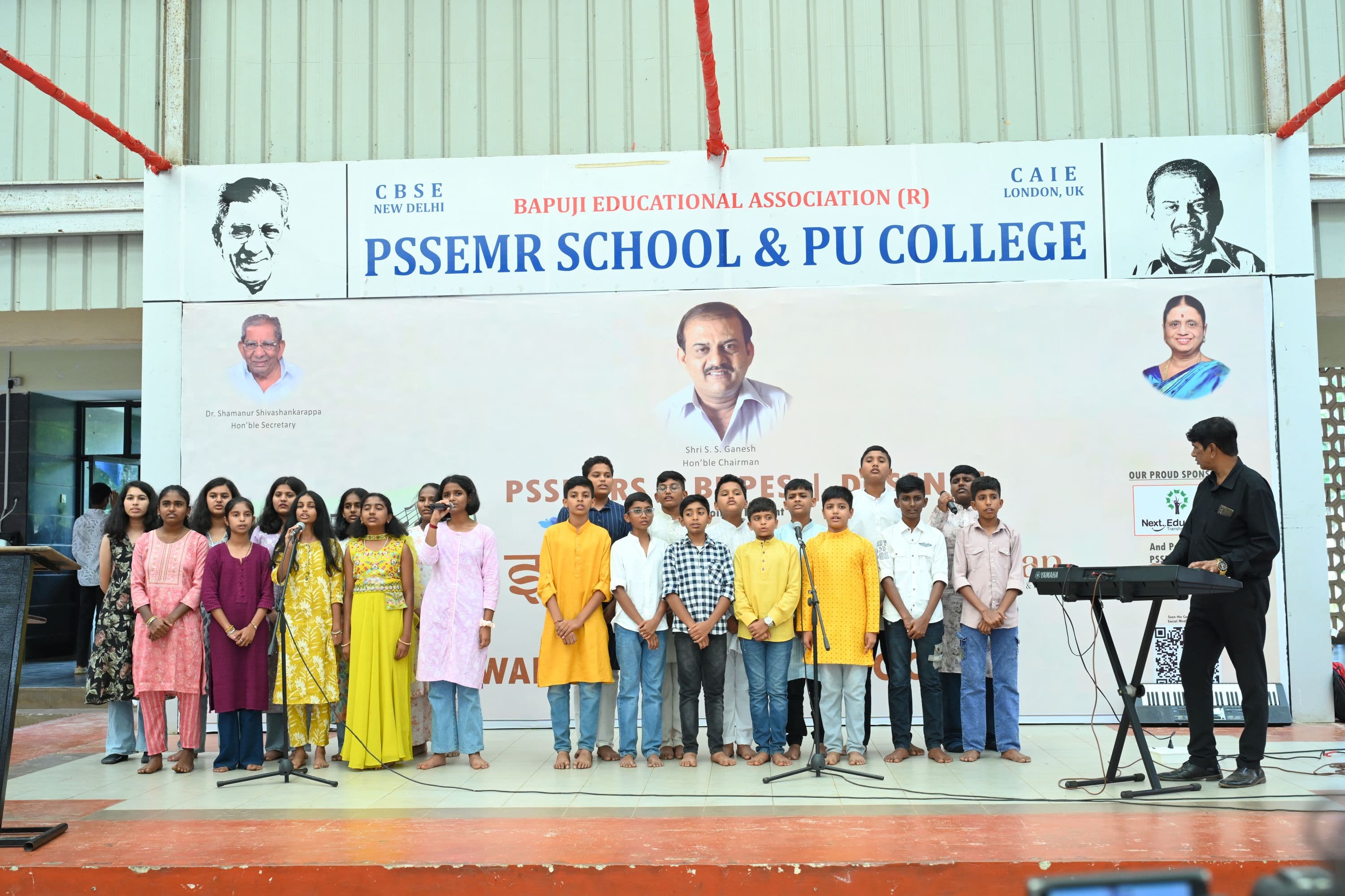 A group of students singing on the assembly-area stage at Sambhram 2K25, beneath a banner naming the host institution and its affiliations.