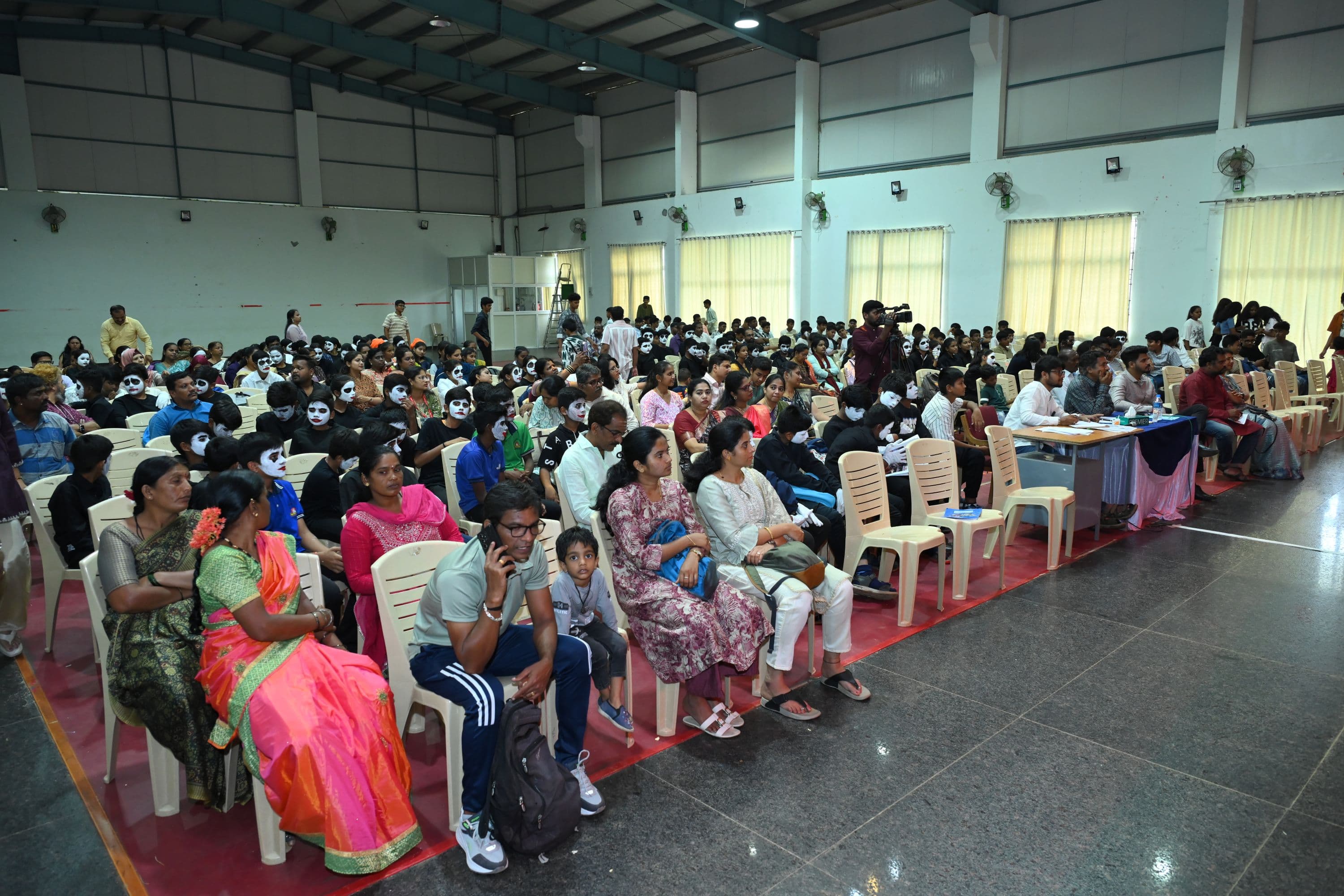 A full indoor hall at Sambhram 2K25, with rows of seated spectators and dozens of entrants in white mime face paint waiting to compete. A judging table with score papers sits at the right.