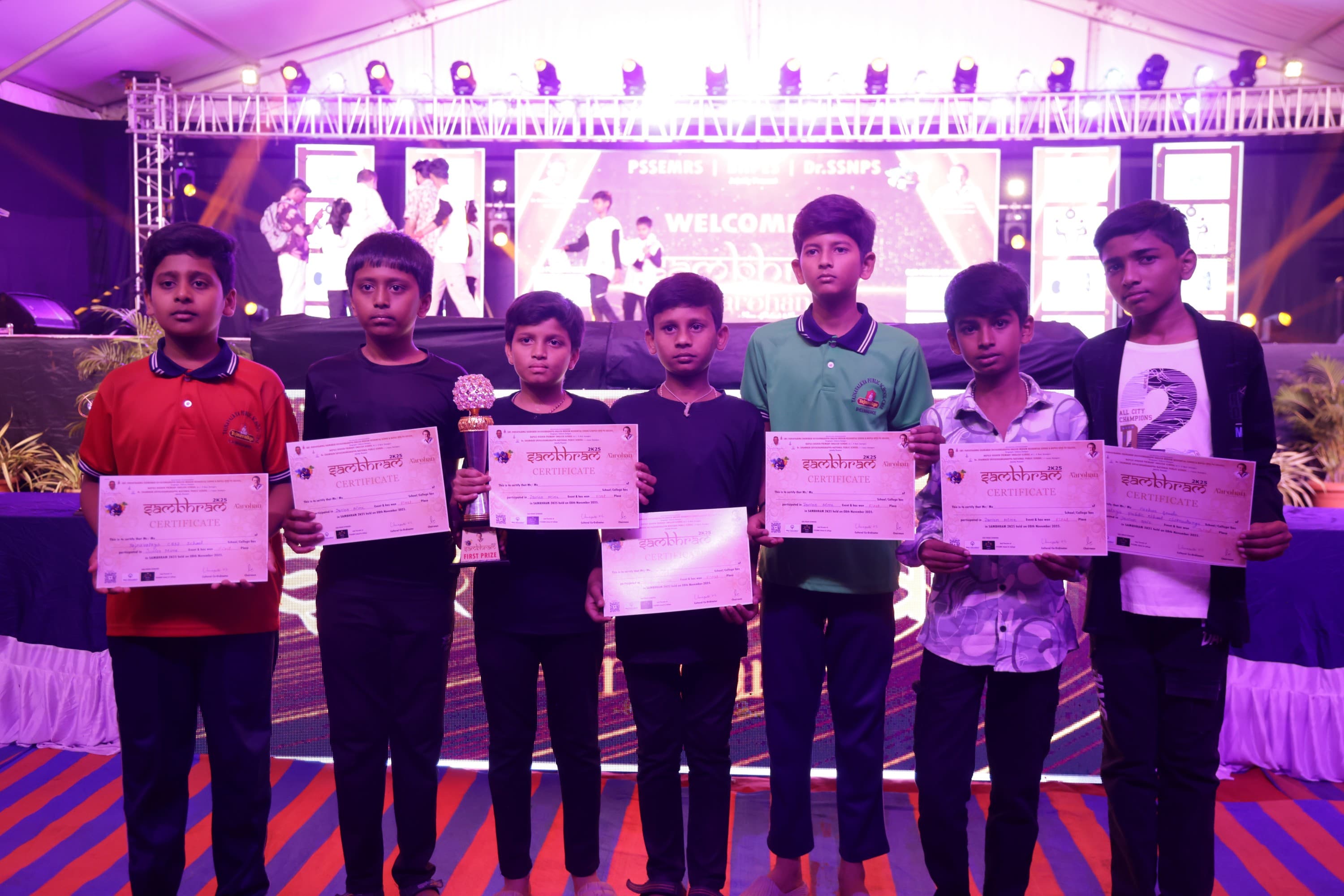 Seven students standing in front of the main stage at Sambhram 2K25, each holding a Sambhram certificate; one also holds a first-prize trophy.