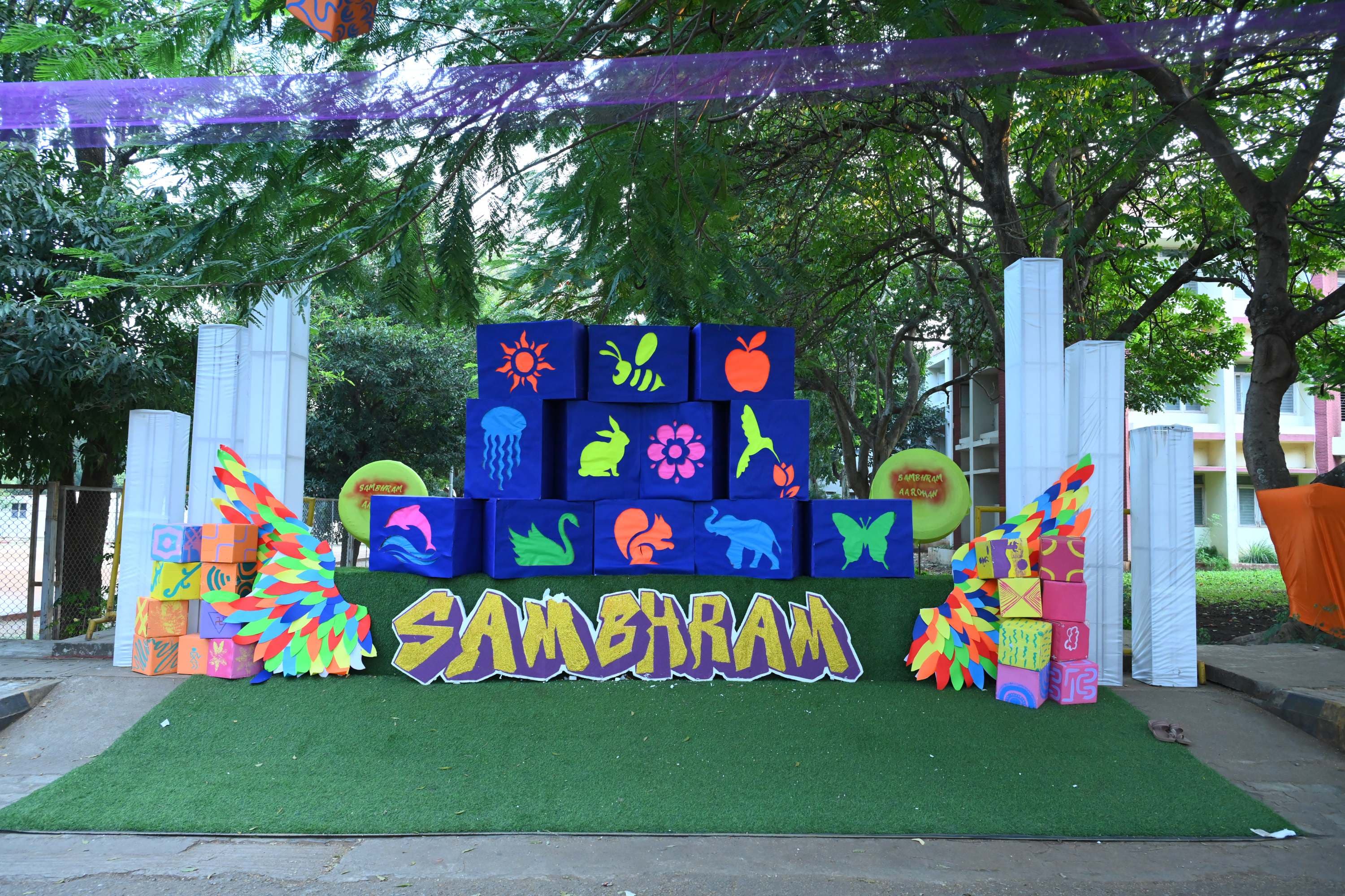 A Sambhram installation on the campus lawn — hand-painted cardboard cubes stacked into a wall of neon animal and nature motifs, with large cut-out letters spelling SAMBHRAM and paper wings on either side.