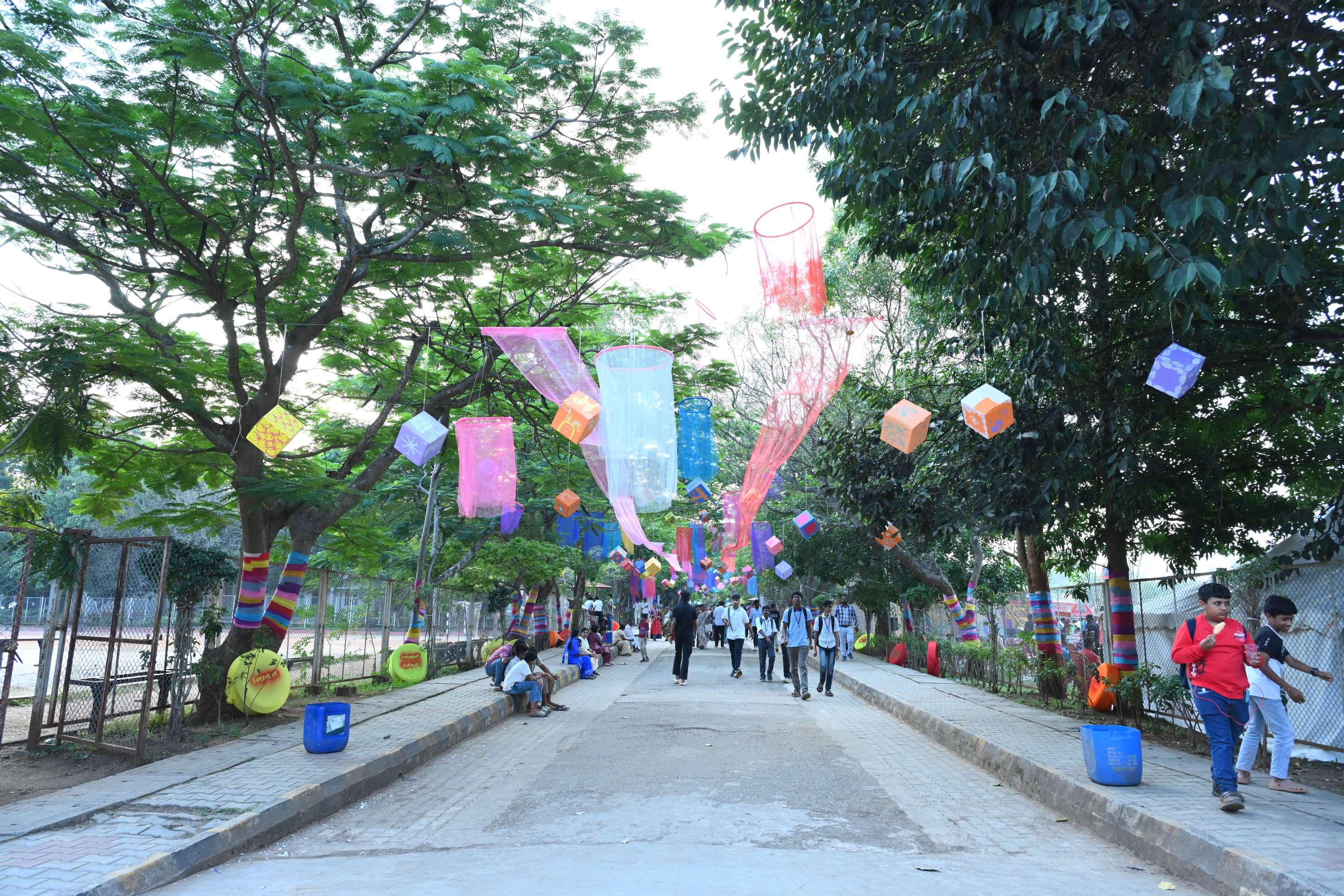 The PSSEMR School campus in Davangere dressed for Sambhram 2K25 — paper lanterns and fabric banners strung between the trees along the approach road as students walk in.