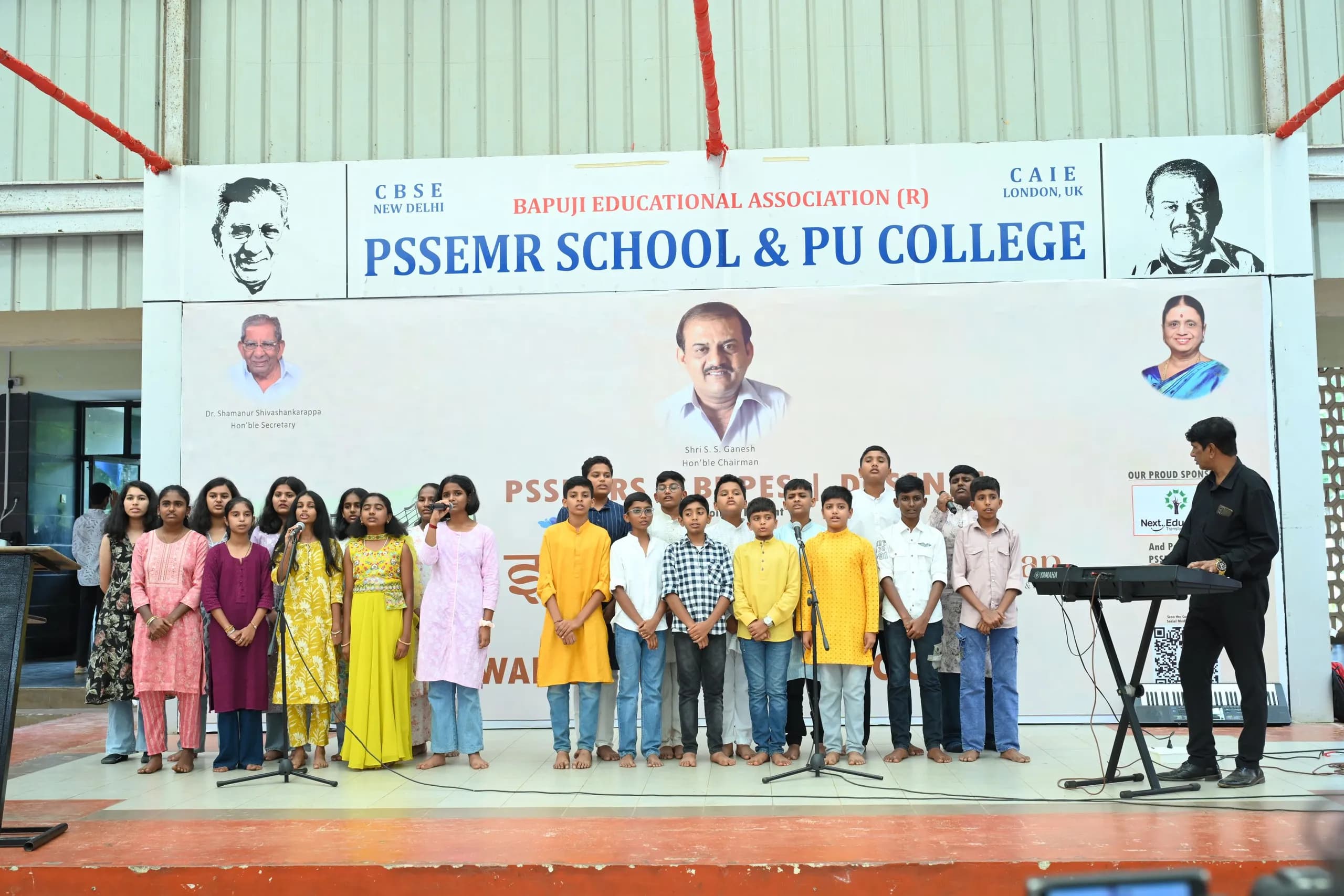 A group of students singing on the assembly-area stage at Sambhram 2K25, beneath a banner naming the host institution and its affiliations.