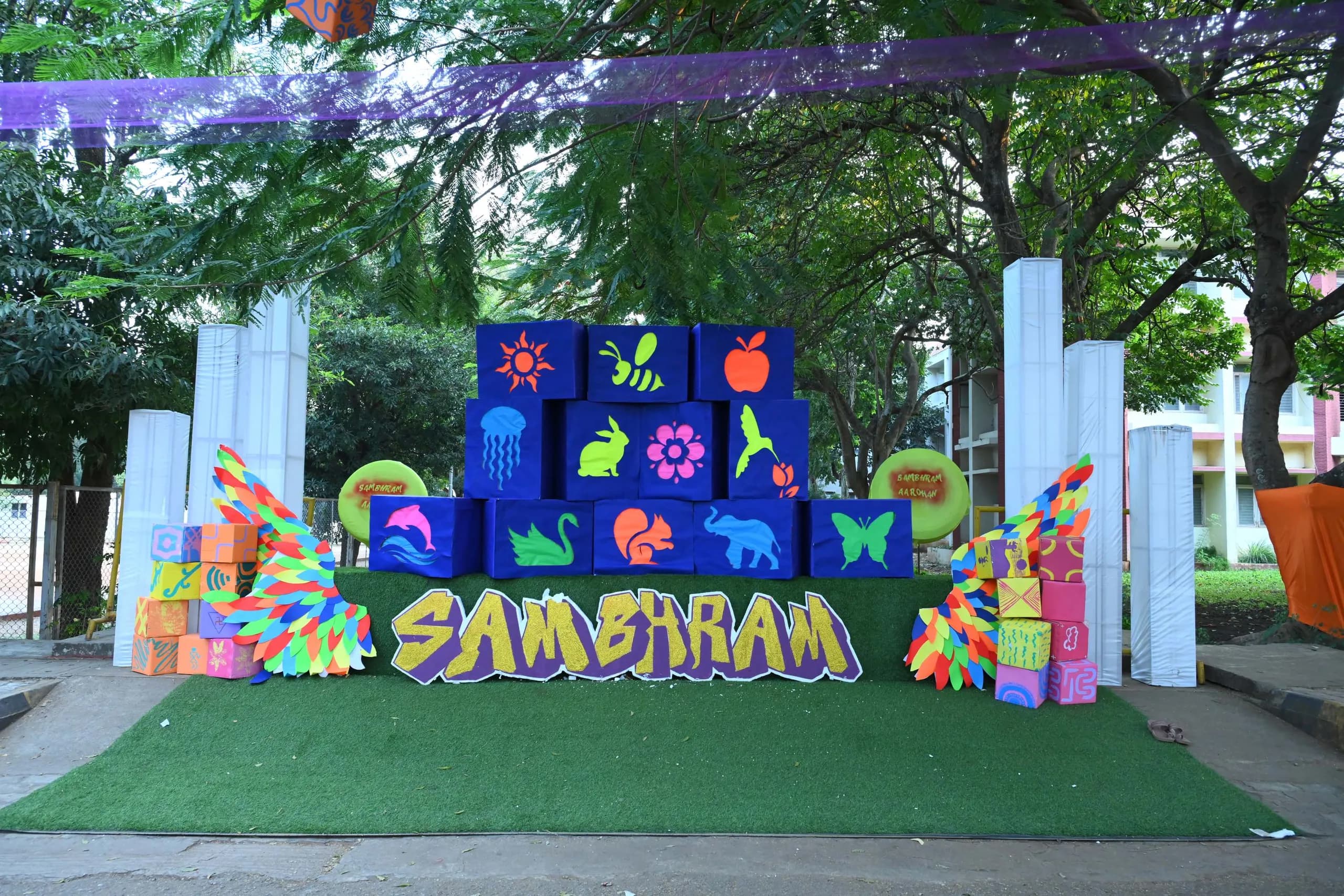 A Sambhram installation on the campus lawn — hand-painted cardboard cubes stacked into a wall of neon animal and nature motifs, with large cut-out letters spelling SAMBHRAM and paper wings on either side.